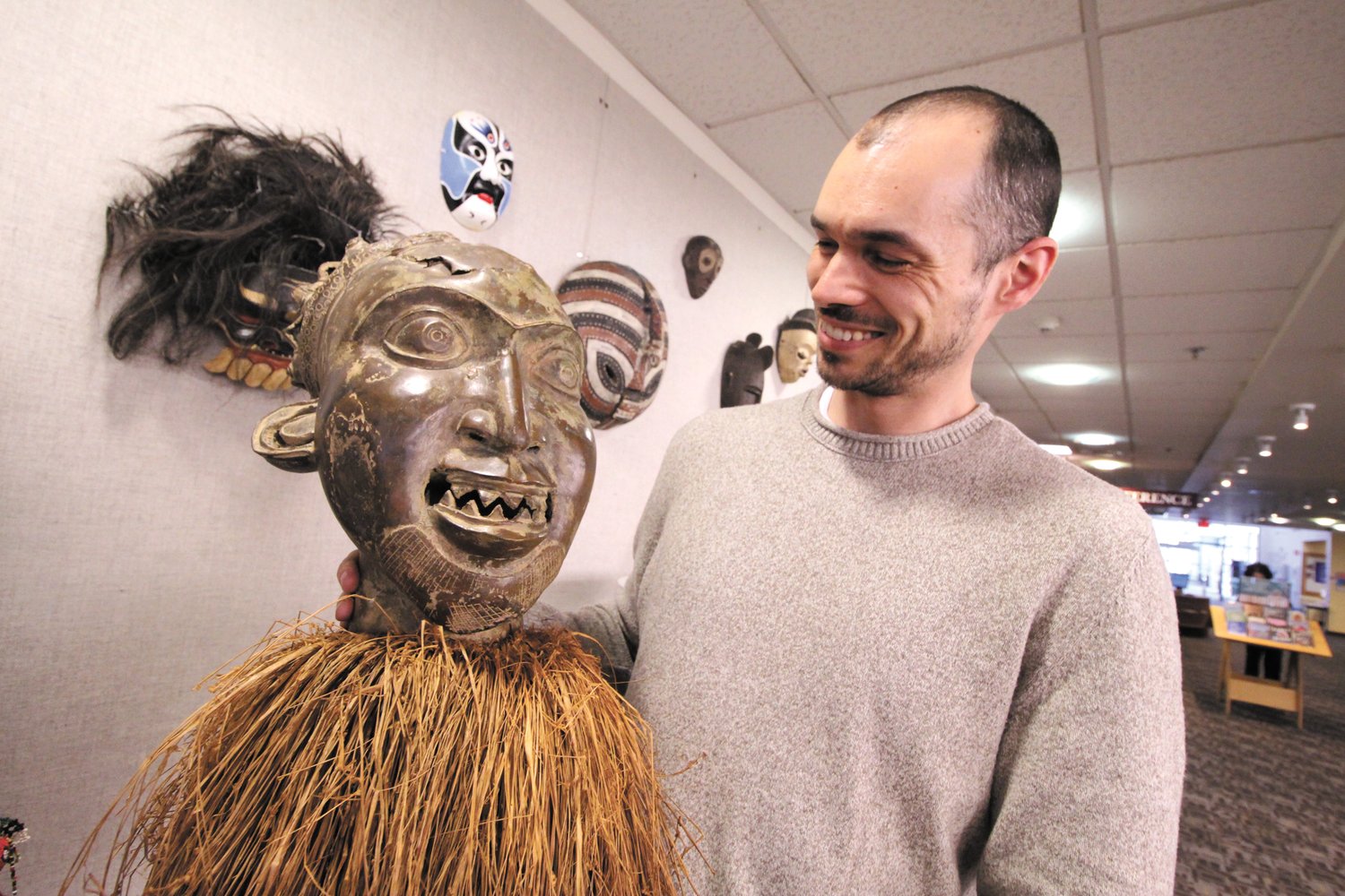FACE TO FACE: Justin Bibee with a Bamum mask from Cameroon, specifically the Cameroon Grasslands, made of bronze with raffia palms. This mask is part of a family of masks that perform together. It represents the male human face and symbolizes a clan ancestor. Masks such as these are performed at funeral or memorial celebrations. This mask is worn on the top of the head, while raffia palm conceals the performer’s face, and a mesh veil covers the performer’s body. It is one of many in his collection on display at the Warwick Public Library through May 31.
