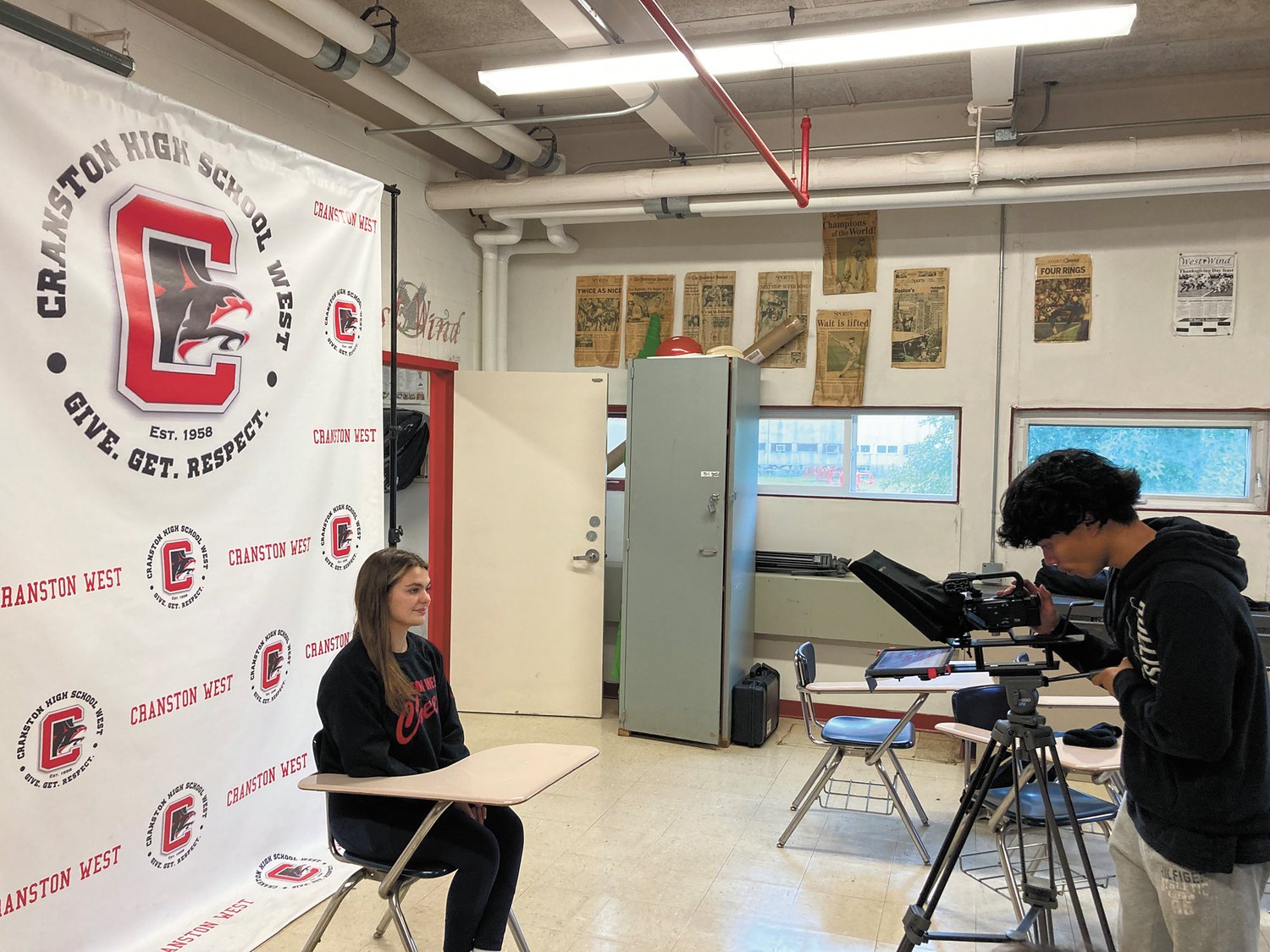 ON AIR: Eric Zhang readies the camera while Gisele Rotolu sits in front of Cranston West’s backdrop gearing up to read from the teleprompter. (Herald photo)